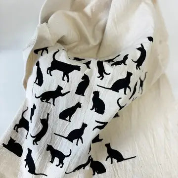 A cream-colored tea towel, featuring a black silhouette pattern of cats in various poses. The fabric is draped elegantly against a neutral light background.