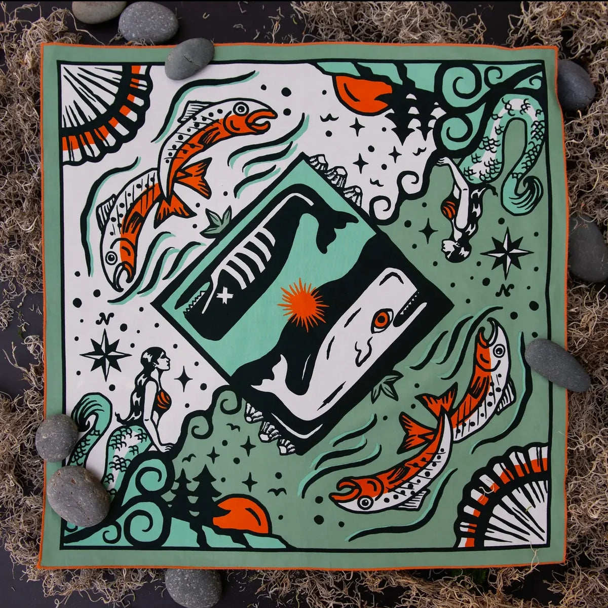 A square bandana with a hand-painted folk art design featuring a central tarot card motif, orange fish, mermaids, and stars on a sage green background, styled flat-lay on dark moss with gray stones.