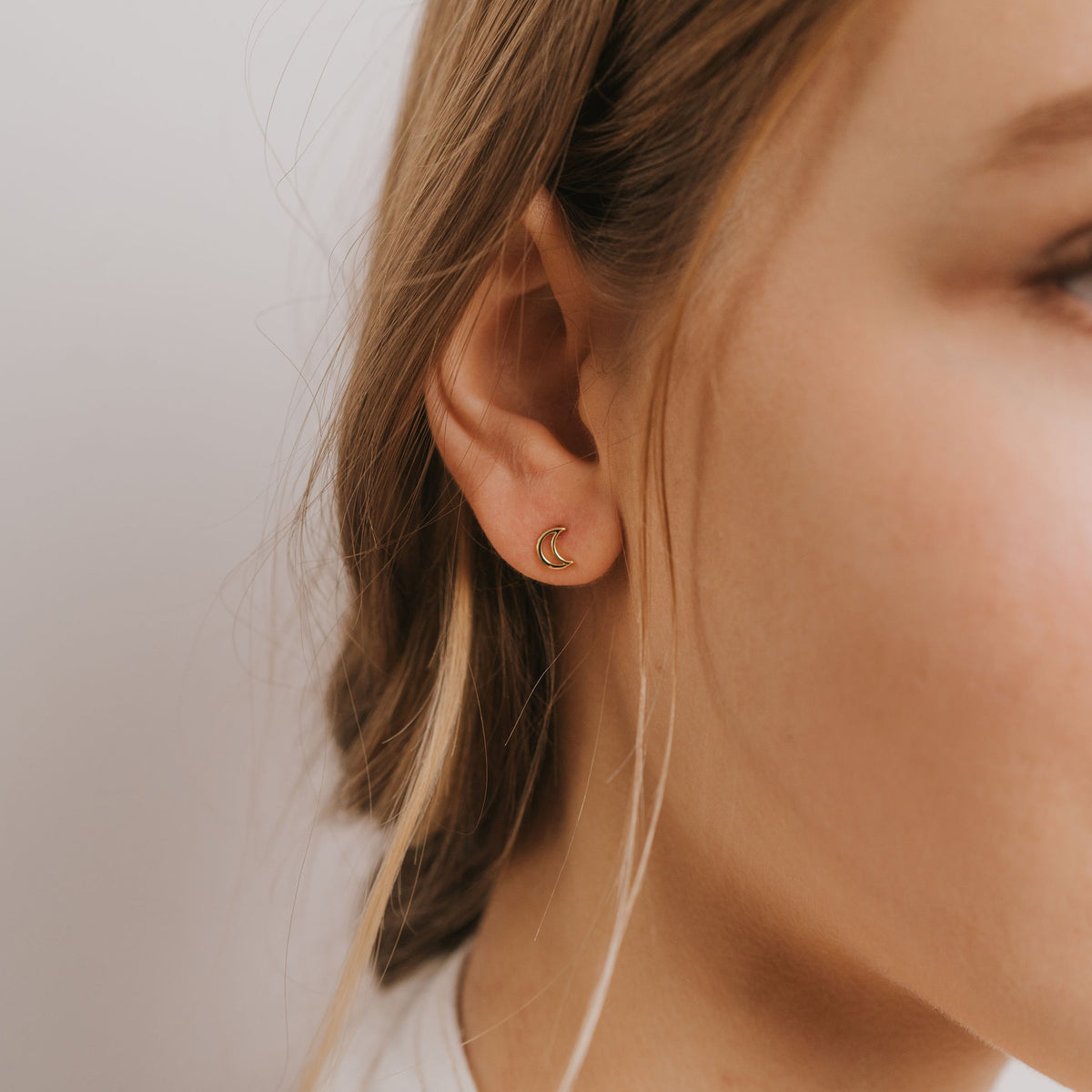 A close-up of a person's ear wearing a small crescent moon stud earring in a warm gold tone against a soft light gray background.