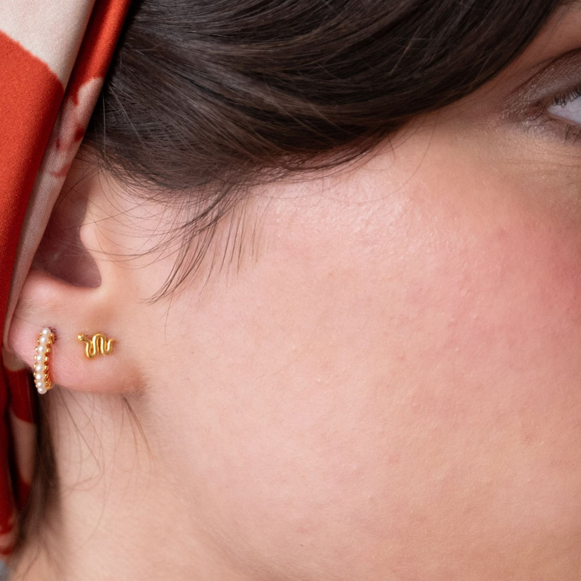 Close-up of a person's ear wearing a small gold snake stud earring and a gold huggie hoop adorned with tiny white pearls, with dark hair and a red-and-white headscarf visible.