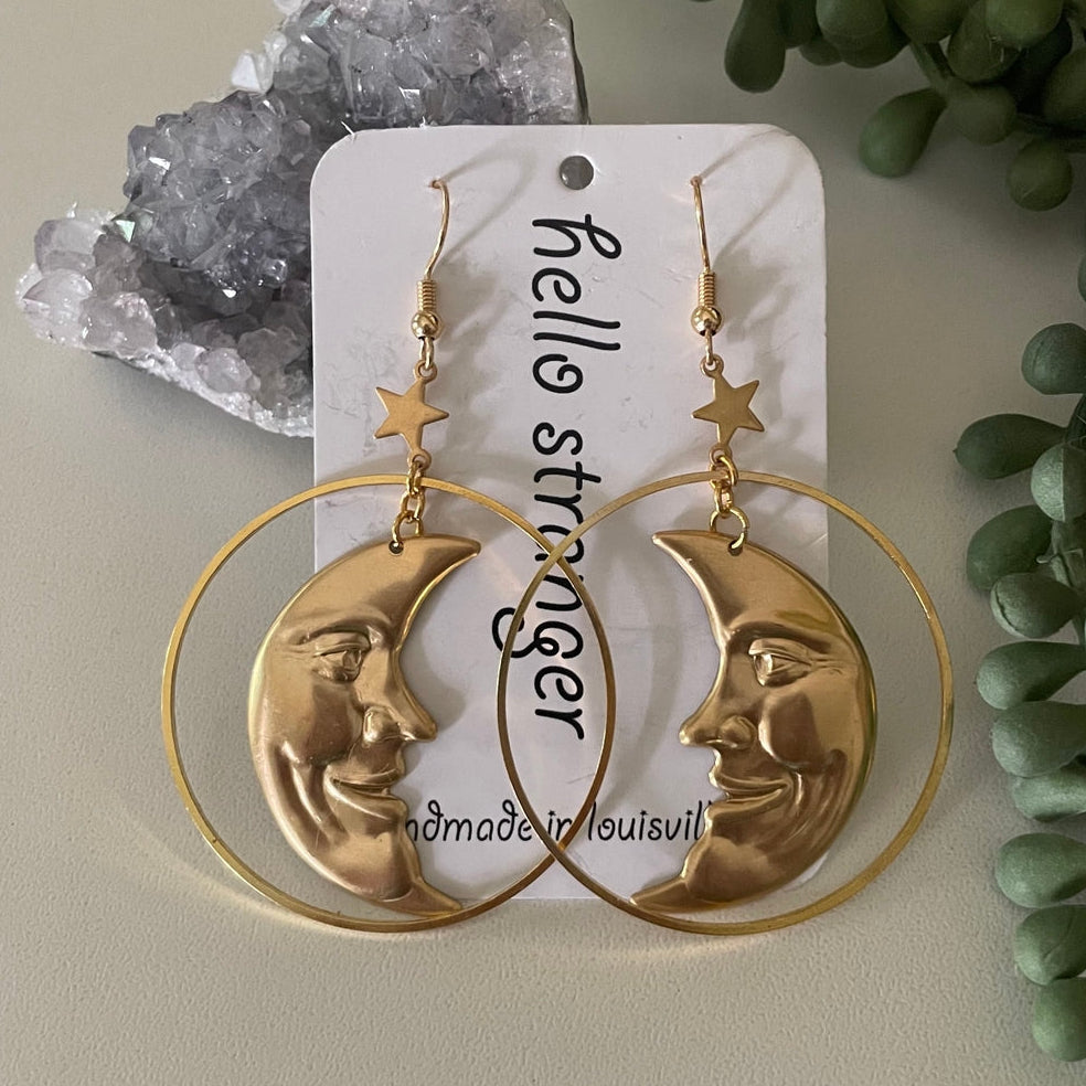 A pair of gold-toned hoop earrings with crescent moon and star motifs, displayed on a white card that reads 'hello stranger' with 'omadein louisville' printed below. The earrings are styled with a crystal cluster and green foliage on a neutral beige background.