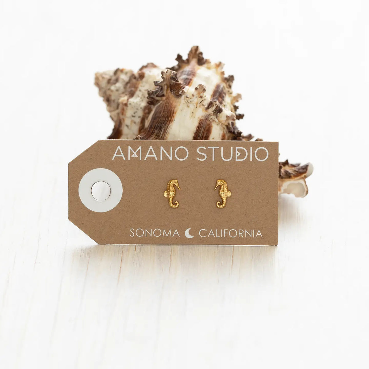 A pair of small gold seahorse stud earrings displayed on a brown cardboard tag that reads 'AMANO STUDIO' and 'SONOMA CALIFORNIA,' with a seashell behind the tag on a white surface.