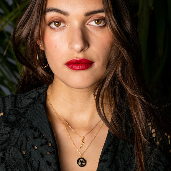 A close-up portrait of a woman with long dark brown hair, wearing layered gold necklaces, one with a black scale of justice pendant, and red lipstick, set against a dark background with green foliage.