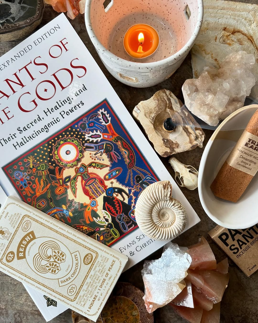 Assorted items on wooden surface: book, chocolate bar in package, lit candle, ceramic items, incense, fossils, skull, crystals