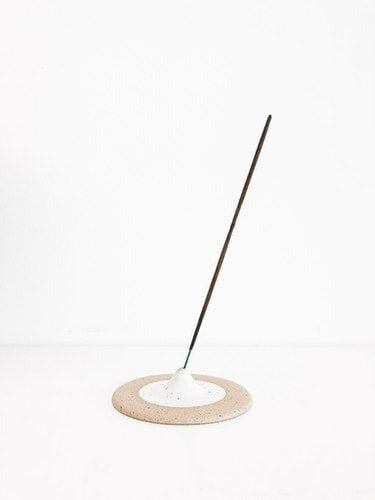 A beige and white incense holder with a circular base holding a single incense stick, placed on a white surface.