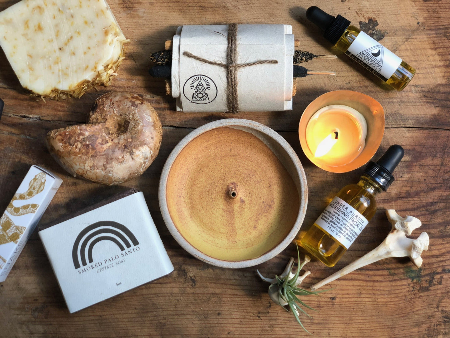A variety of items including soap, an ammonite, a dish for burning, incence sticks, candle, crystals, a plant, a bone, home decor, bath and body products, all laid out on a wooden surface.