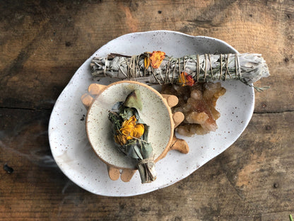 Sage bundled with flowers burning on a white dish, on top of another larger white dish with a crystal and large sage bundle, all on a wooden surface