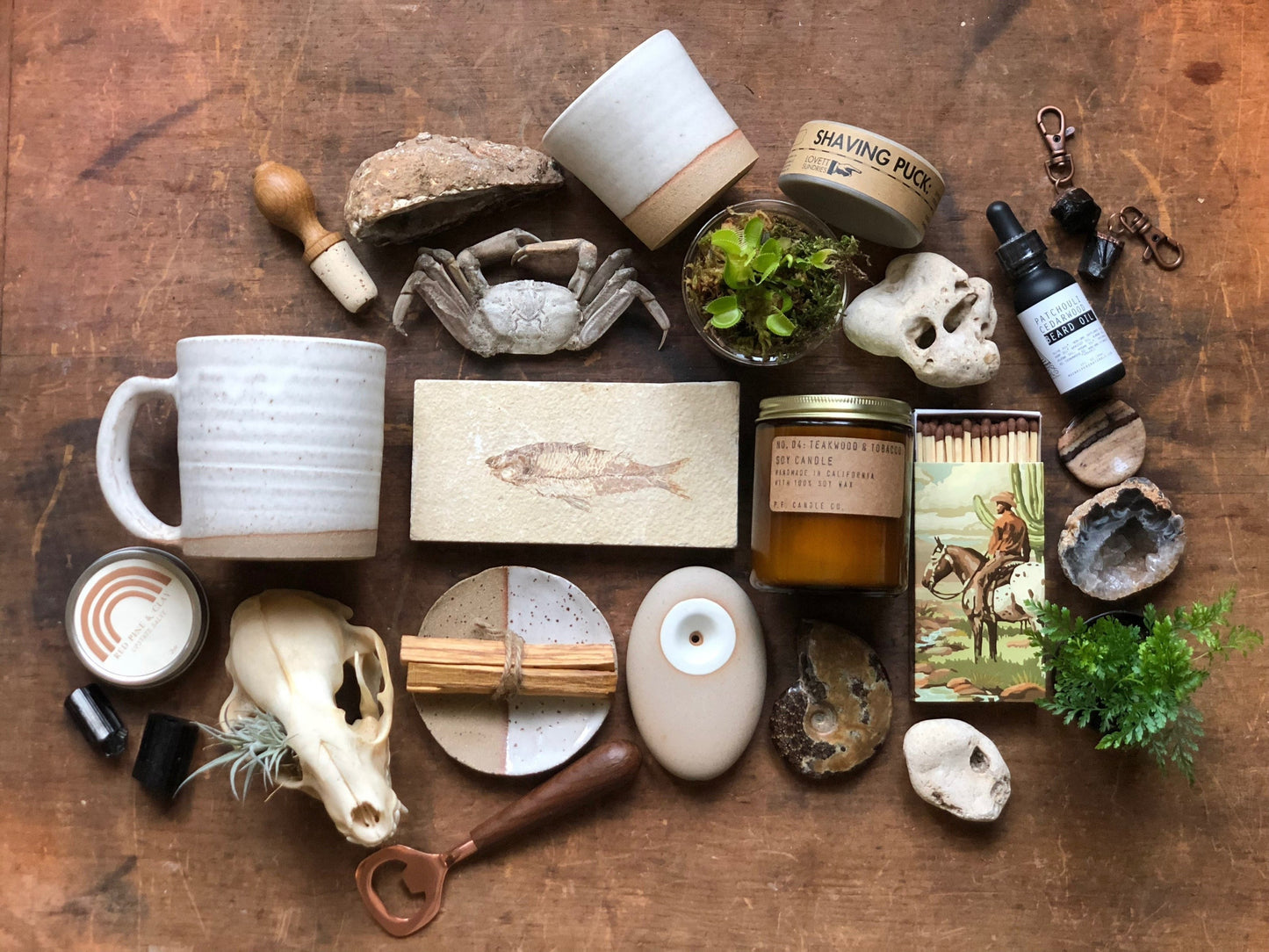 A variety of items including Palo Santo wood, white sage, a dish for burning, crystals, a plant, home decor, bath and body products, ceramics, a sheepskin rug, and a book, all laid out on a wooden surface.