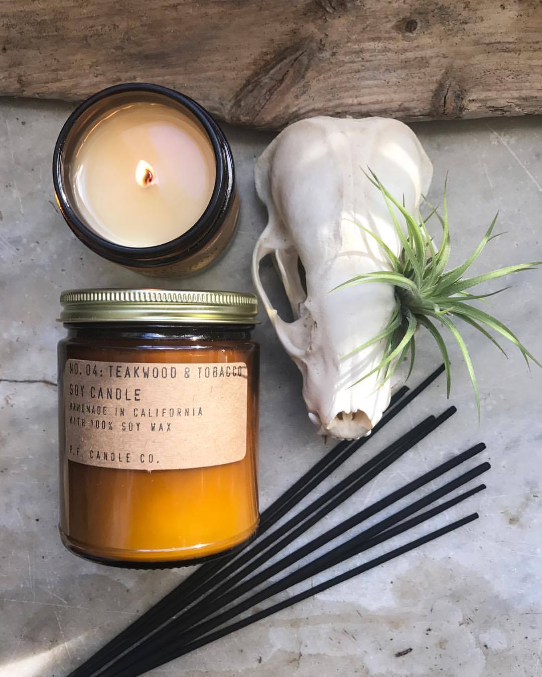 An amber glass jar candle with a teakwood and tobacco label, a lit candle, a skull, and incense sticks on a concrete background.
