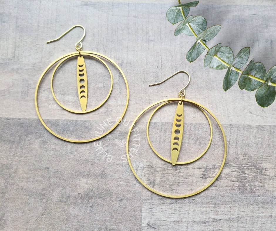 A pair of gold-toned hoop earrings with a central vertical disc featuring crescent moon cutouts, displayed on a gray wood-textured background with a green eucalyptus sprig in the corner.