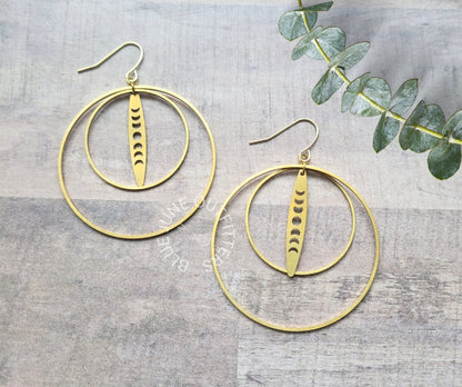 A pair of gold-toned hoop earrings with a central vertical disc featuring crescent moon cutouts, displayed on a gray wood-textured background with a green eucalyptus sprig in the corner.