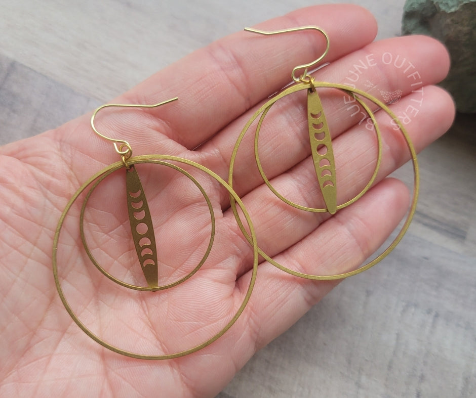 Gold-toned double hoop earrings with a dangling vertical disc featuring moon phase cutouts, shown resting on an open hand.