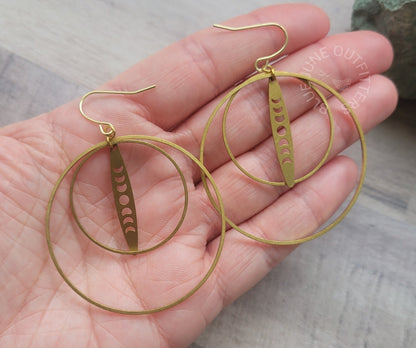 Gold-toned double hoop earrings with a dangling vertical disc featuring moon phase cutouts, shown resting on an open hand.