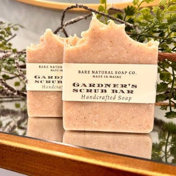 Two Gardner's Scrub Bars from Bare Natural Soap Co. on a reflective surface with greenery in the background.