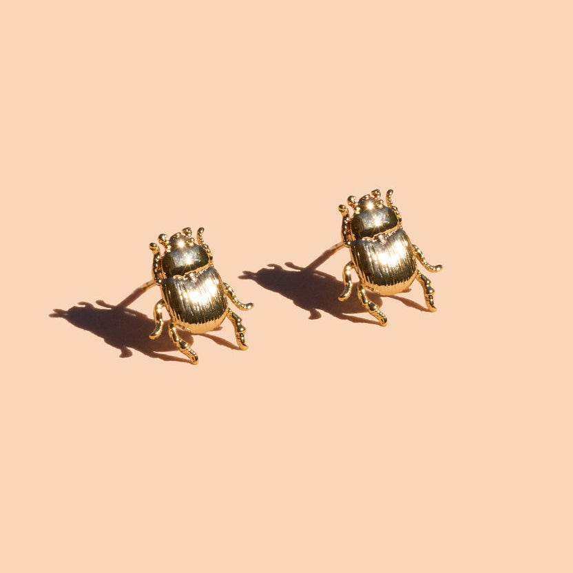 A pair of gold beetle stud earrings on a solid peach background with soft shadows.