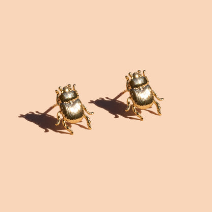 A pair of gold beetle stud earrings on a solid peach background with soft shadows.