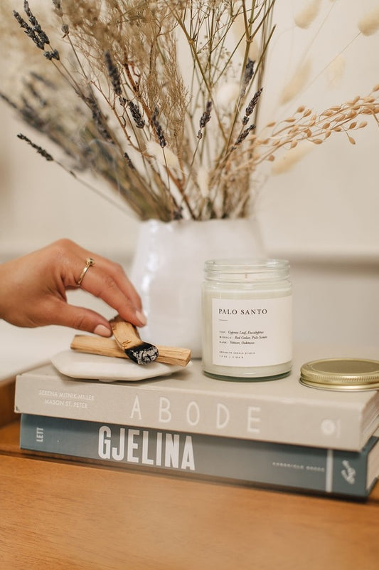 Person lighting a Palo Santo incense stick next to a candle and books on a wooden surface.