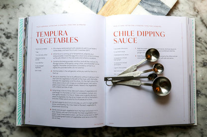 Open cookbook with recipes for tempura vegetables and chile dipping sauce, with measuring spoons on top.