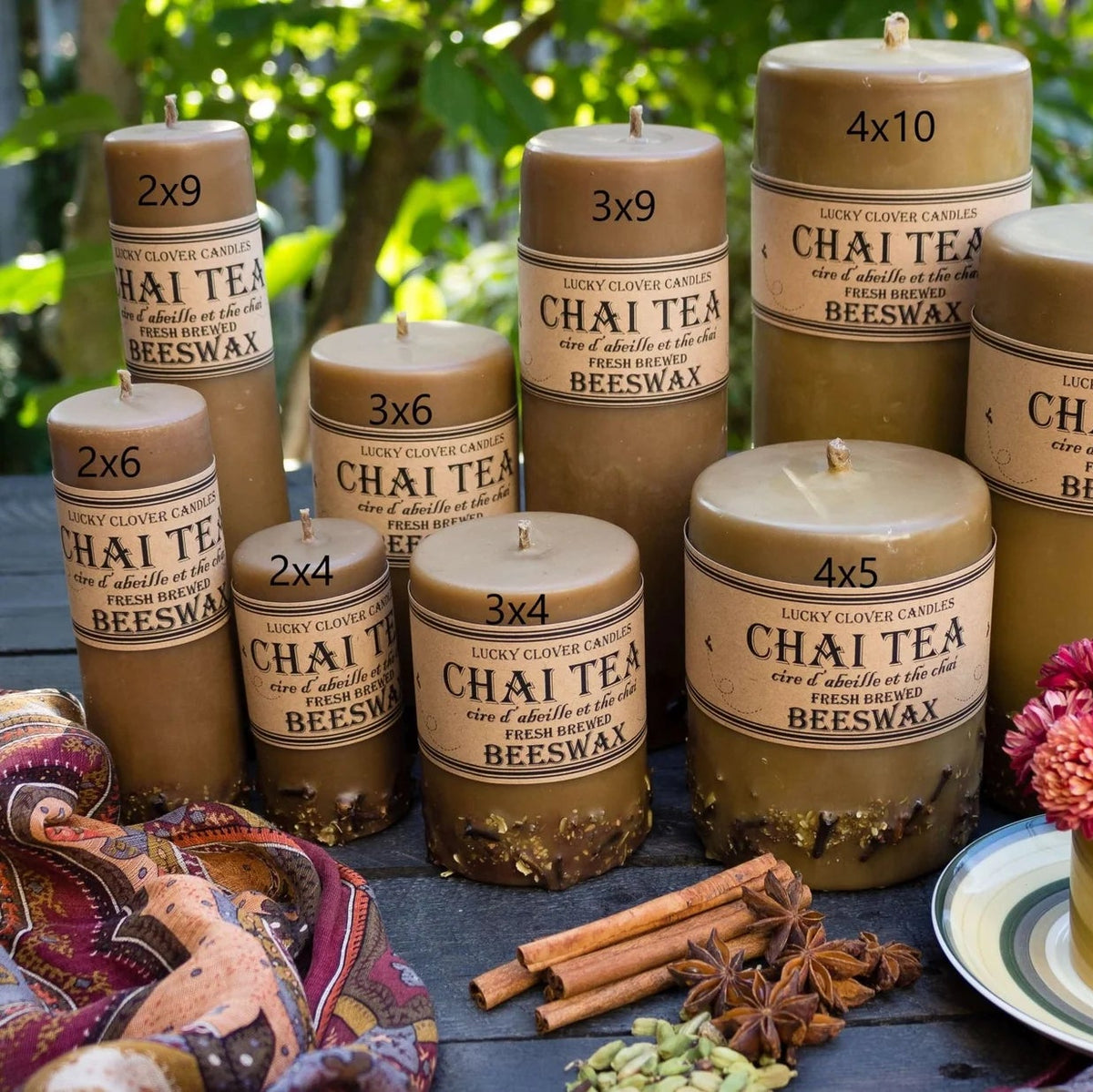Chai tea candles with labels on a wooden table outdoors, surrounded by flowers and spices.