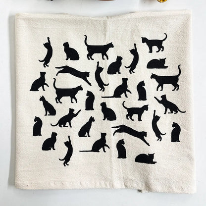 An off-white fabric tea towel featuring a collection of black cat silhouettes in various poses, laid flat on a white surface.