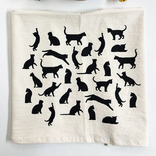An off-white fabric tea towel featuring a collection of black cat silhouettes in various poses, laid flat on a white surface.