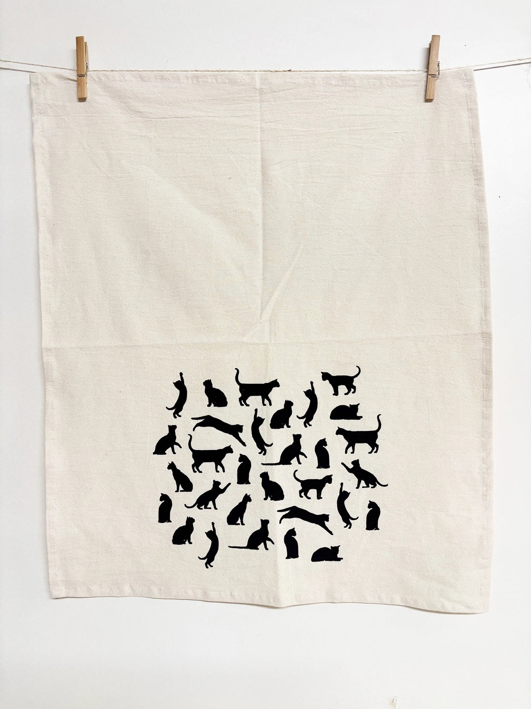 An off-white linen tea towel with a black cat silhouette pattern, hanging from a clothesline with wooden clips against a white wall.