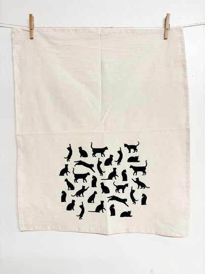 An off-white linen tea towel with a black cat silhouette pattern, hanging from a clothesline with wooden clips against a white wall.
