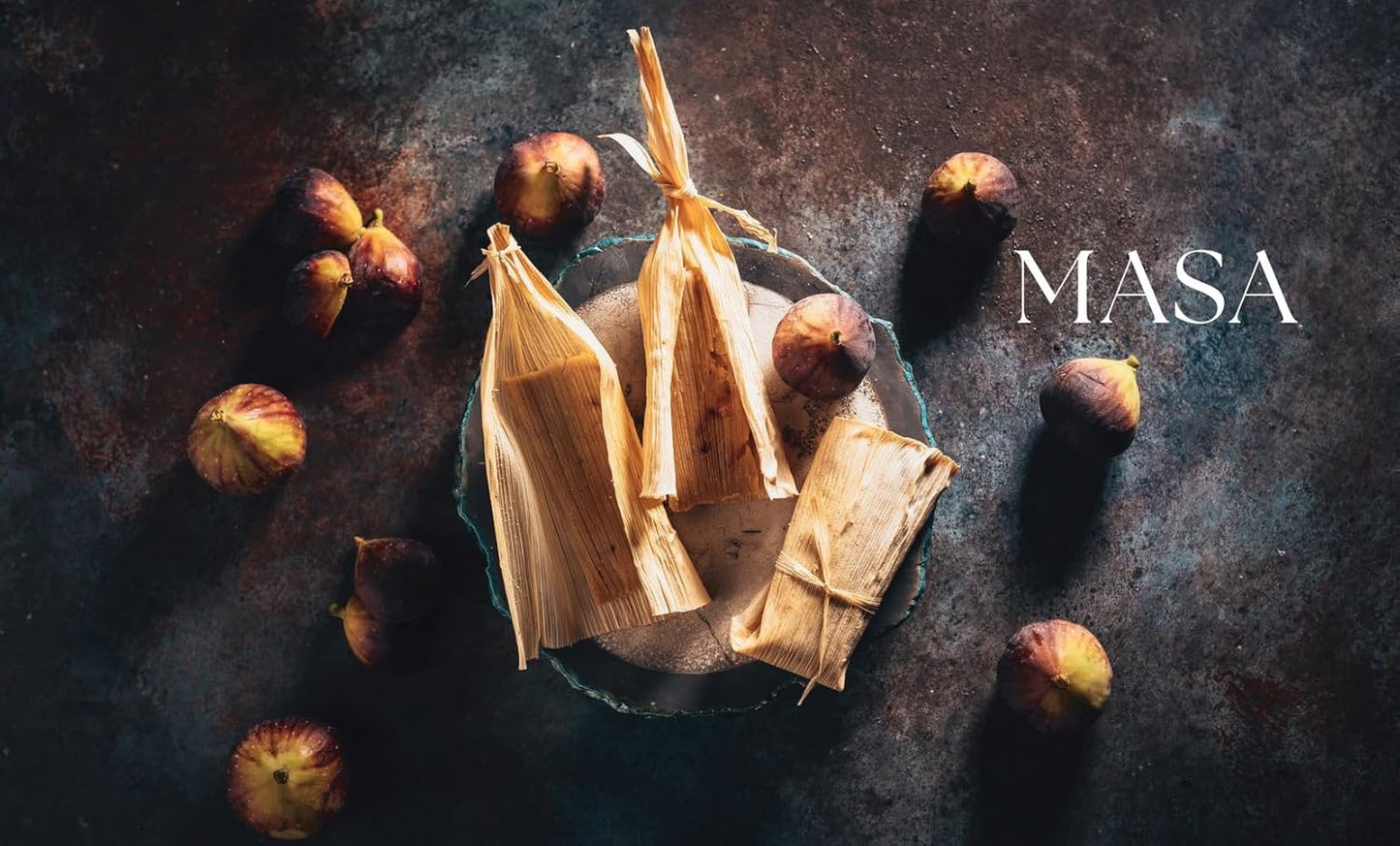 Tamales wrapped in corn husks with figs on a dark surface, featuring the brand 'MASA'.