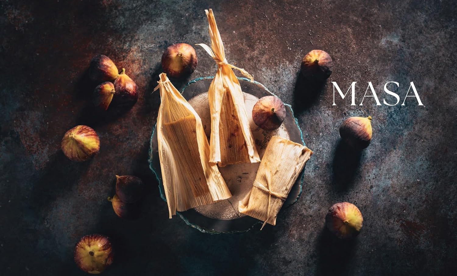 Tamales wrapped in corn husks with figs on a dark surface, featuring the brand 'MASA'.