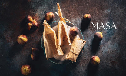 Tamales wrapped in corn husks with figs on a dark surface, featuring the brand 'MASA'.