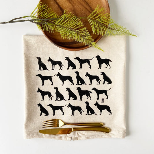 A flat-lay of a cream-colored kitchen towel printed with black dog silhouettes, styled with a wooden plate, pine branches, and gold-toned flatware on a white background.