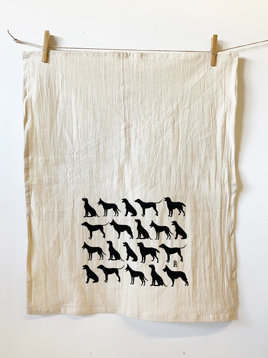 A cream-colored tea towel with black dog silhouettes, hanging from a string with wooden clothespins against a white wall.