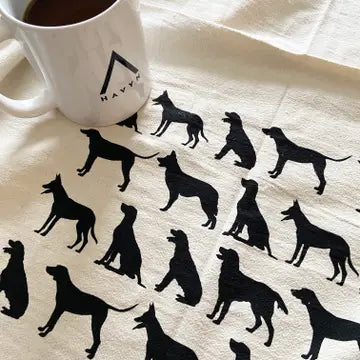 A close-up of a cream-colored tea towel printed with repeated black dog silhouettes, with part of a white mug visible in the upper left corner.