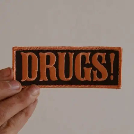 Patch with 'DRUGS!' text held by a hand against a plain background