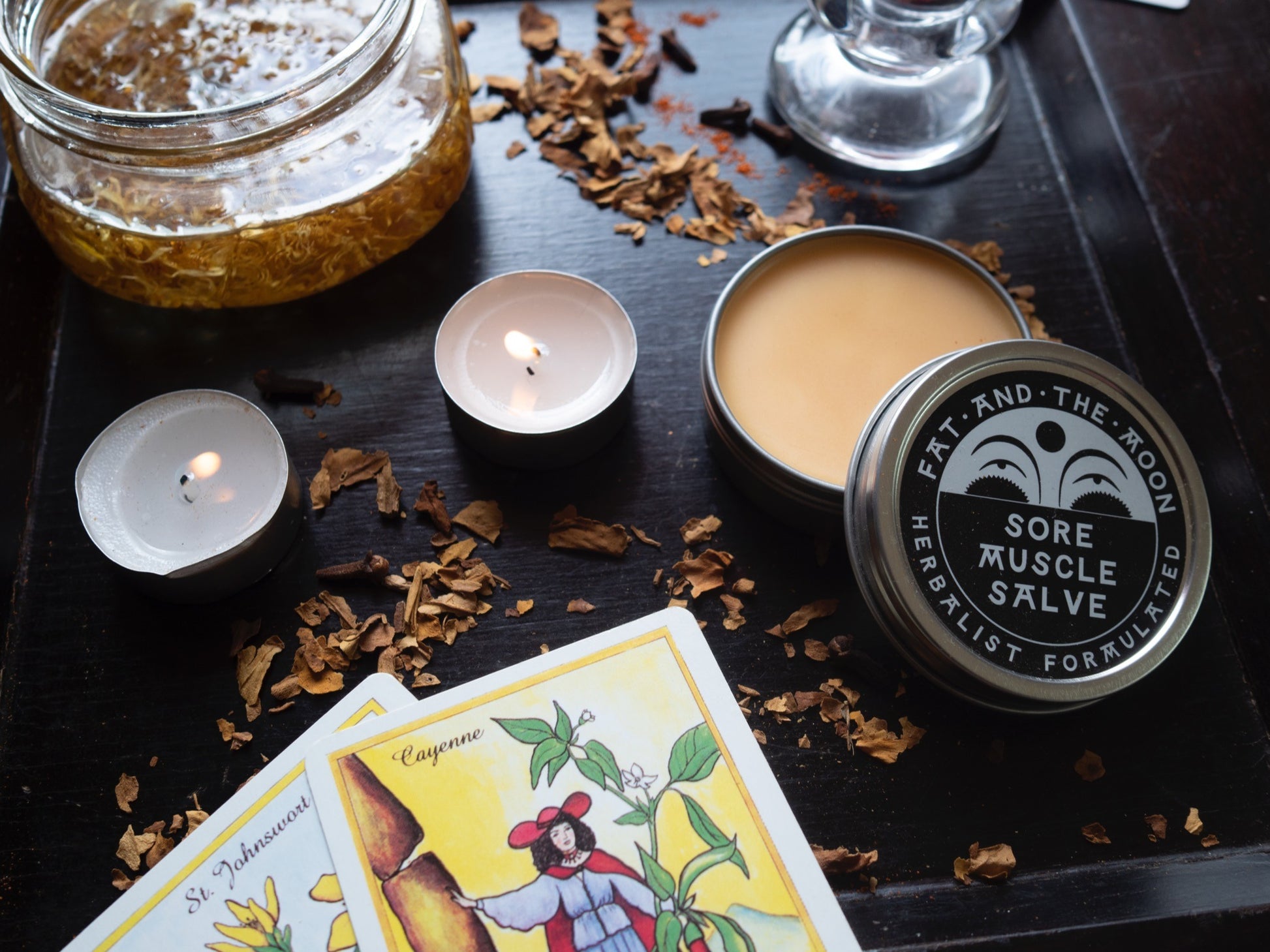 Sore muscle salve tin with candles and tarot cards on a dark surface