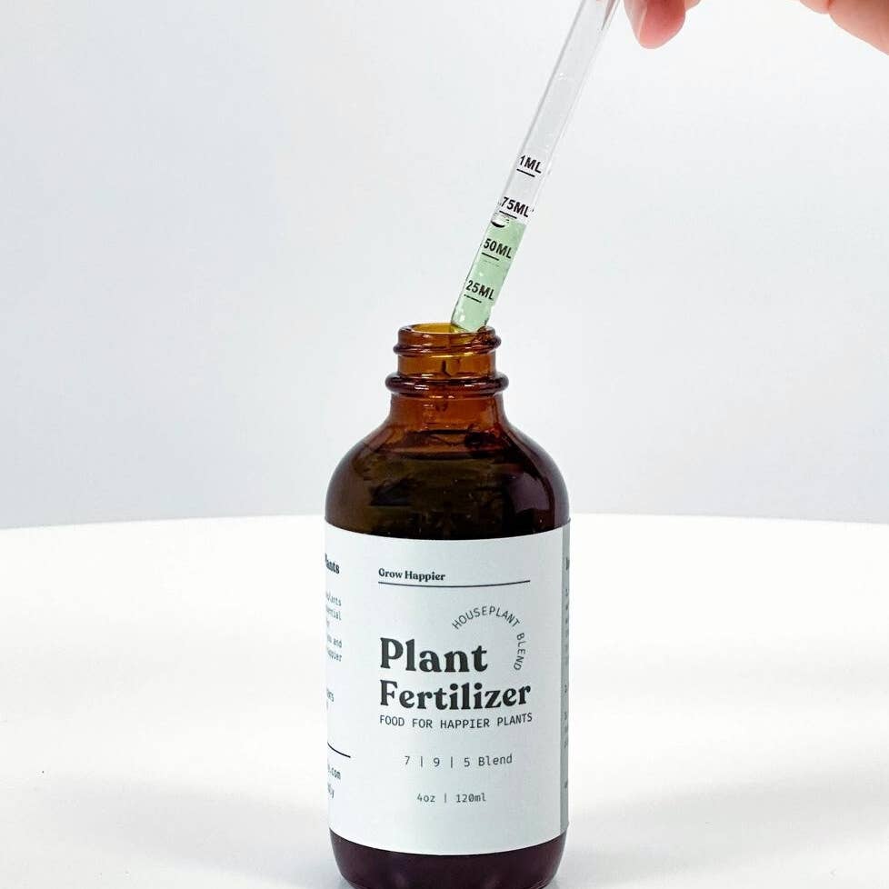 An amber glass bottle of Plant Fertilizer with a white label is shown on a white surface, with a hand holding a glass dropper above the bottle.