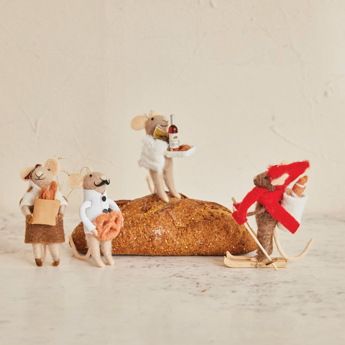 Four small fabric mouse figurines posed around a textured loaf of bread on a light beige surface against a matching neutral background. One mouse wears white and holds a bottle and plate, one stands on the loaf wearing white, one holds a brown bag and orange item, and one wears a red winter outfit with white pants and wooden skis.