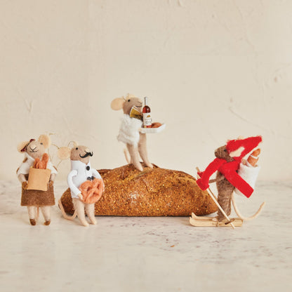 Four small fabric mouse figurines posed around a textured loaf of bread on a light beige surface against a matching neutral background. One mouse wears white and holds a bottle and plate, one stands on the loaf wearing white, one holds a brown bag and orange item, and one wears a red winter outfit with white pants and wooden skis.