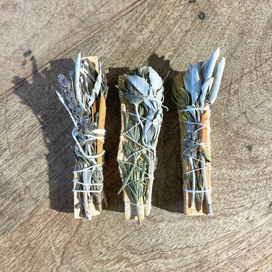 Three bundles of dried herbs and palo santo tied with twine on a wooden surface