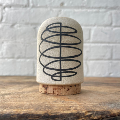Ceramic stash with a black spiral design on a cork base, against a white brick wall.