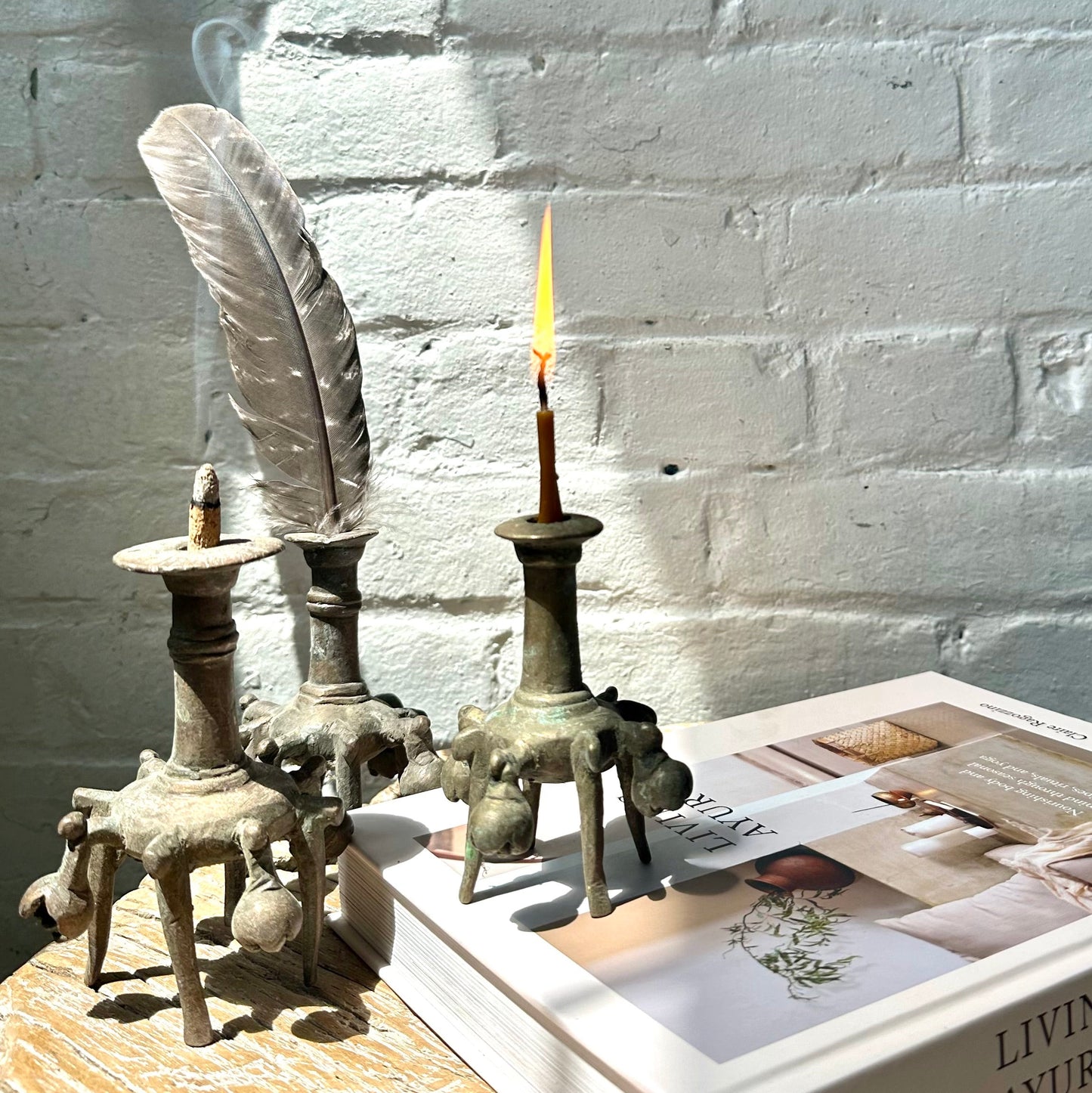 Brass inkwells with lit candle, incense and feather, on top of wooden table, with book, against a white brick background