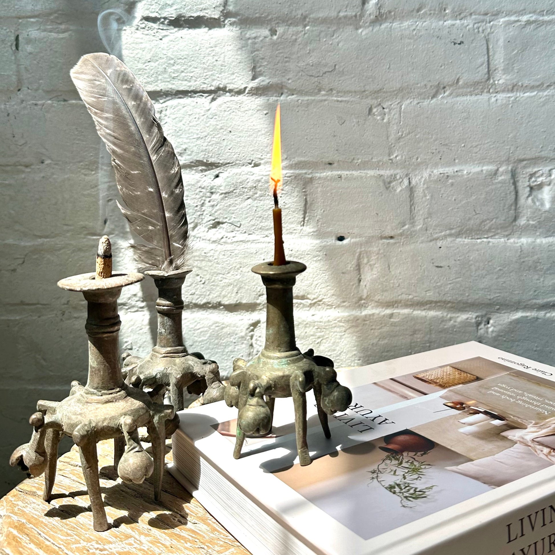 Brass inkwells with lit candle, incense and feather, on top of wooden table, with book, against a white brick background