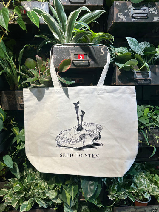 White tote bag with black design and text 'Seed to Stem' on a background of plants.