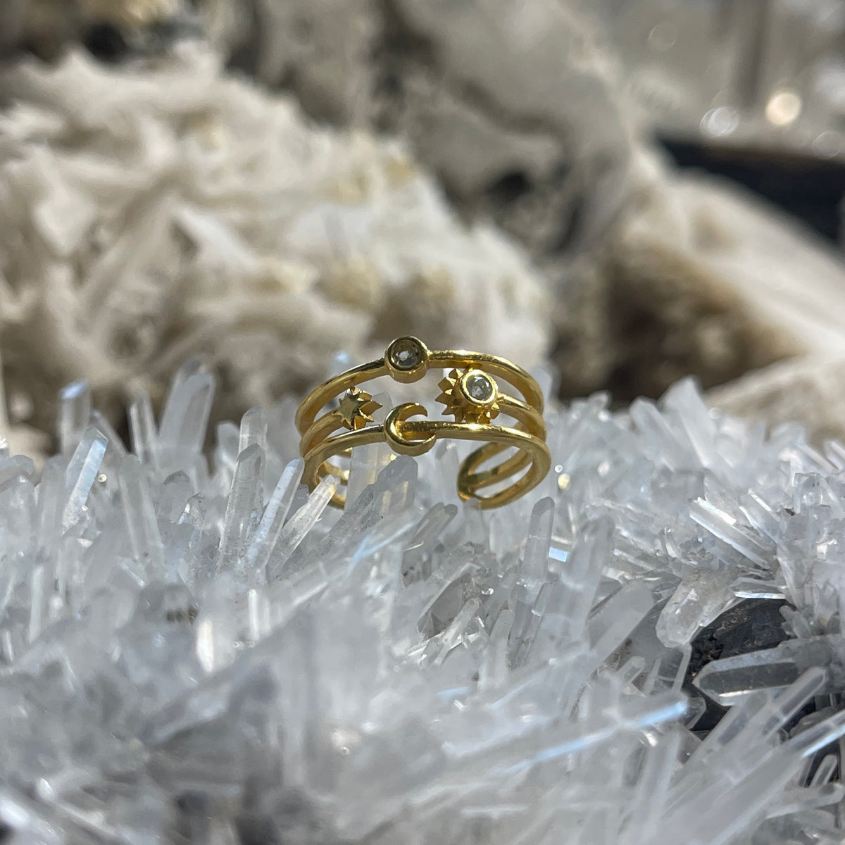 A gold-toned open wrap ring with crescent moon, sun, and small oval details, displayed on a cluster of clear crystals with a blurred white crystal background.