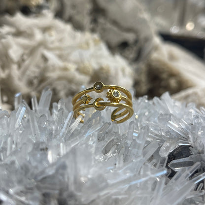 A gold-toned open wrap ring with crescent moon, sun, and small oval details, displayed on a cluster of clear crystals with a blurred white crystal background.