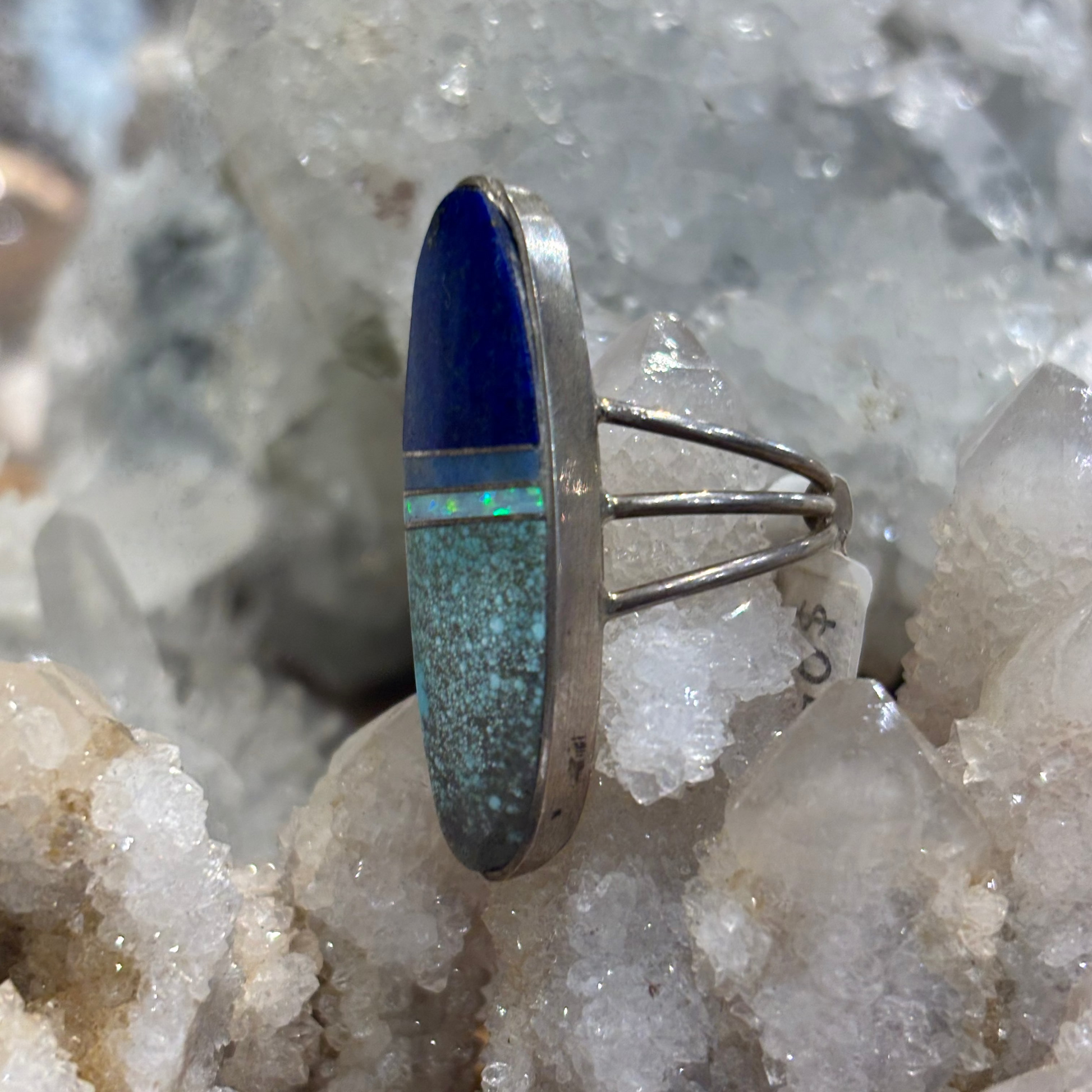 A silver ring featuring a vertical oval inlay with a deep blue top section and a turquoise-blue bottom section, displayed on a bed of white crystal clusters.