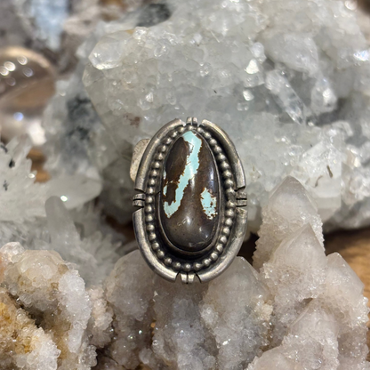 A silver ring featuring an oval cabochon stone with a dark brown base and a bright turquoise-blue matrix, displayed on a cluster of white quartz crystals.