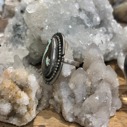 A silver-toned ring with a round turquoise and dark brown stone inlay, displayed on a cluster of white quartz crystals over a wooden surface.