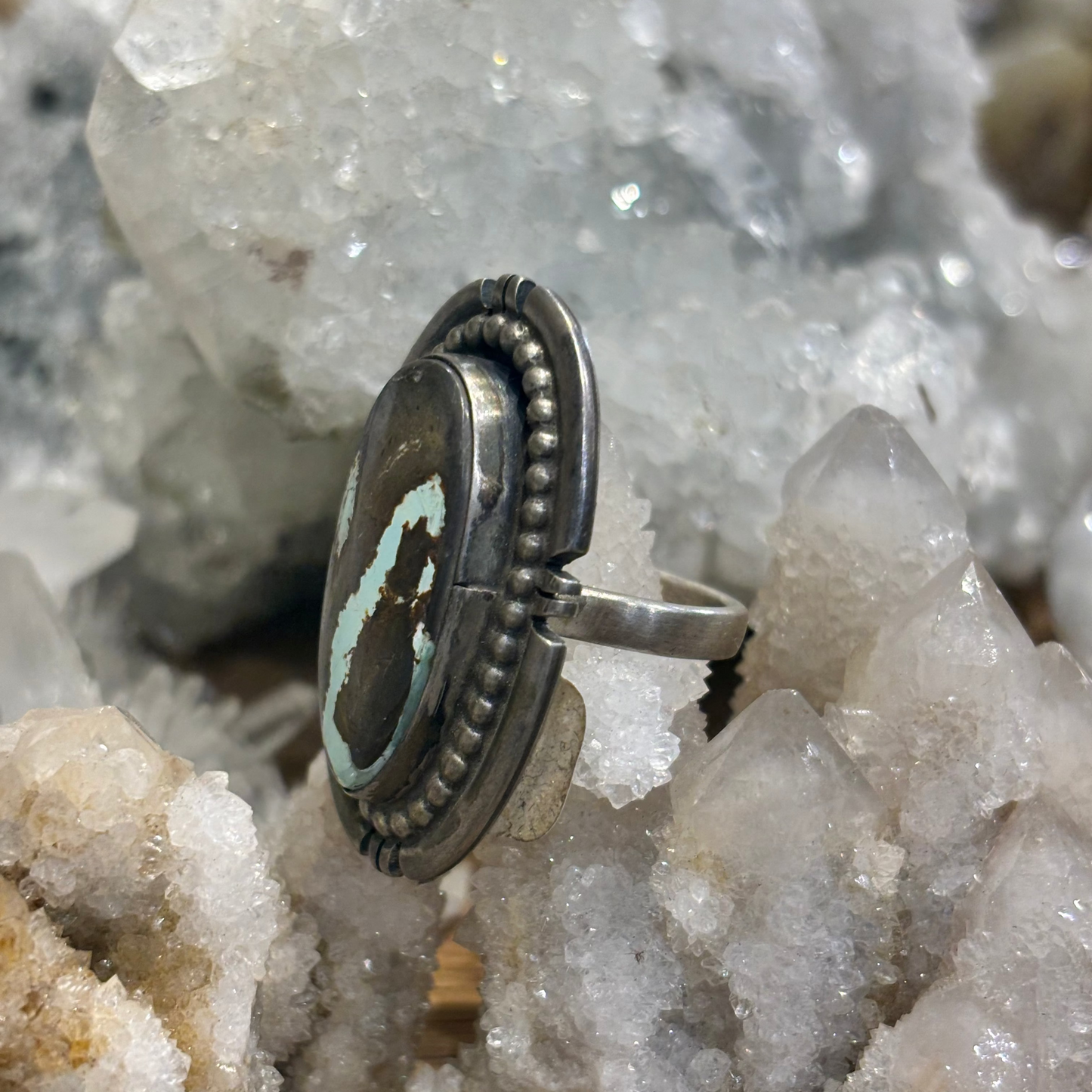 A silver-toned ring with a round dark stone featuring a light turquoise-blue curved inlay, displayed among white and beige crystal clusters.