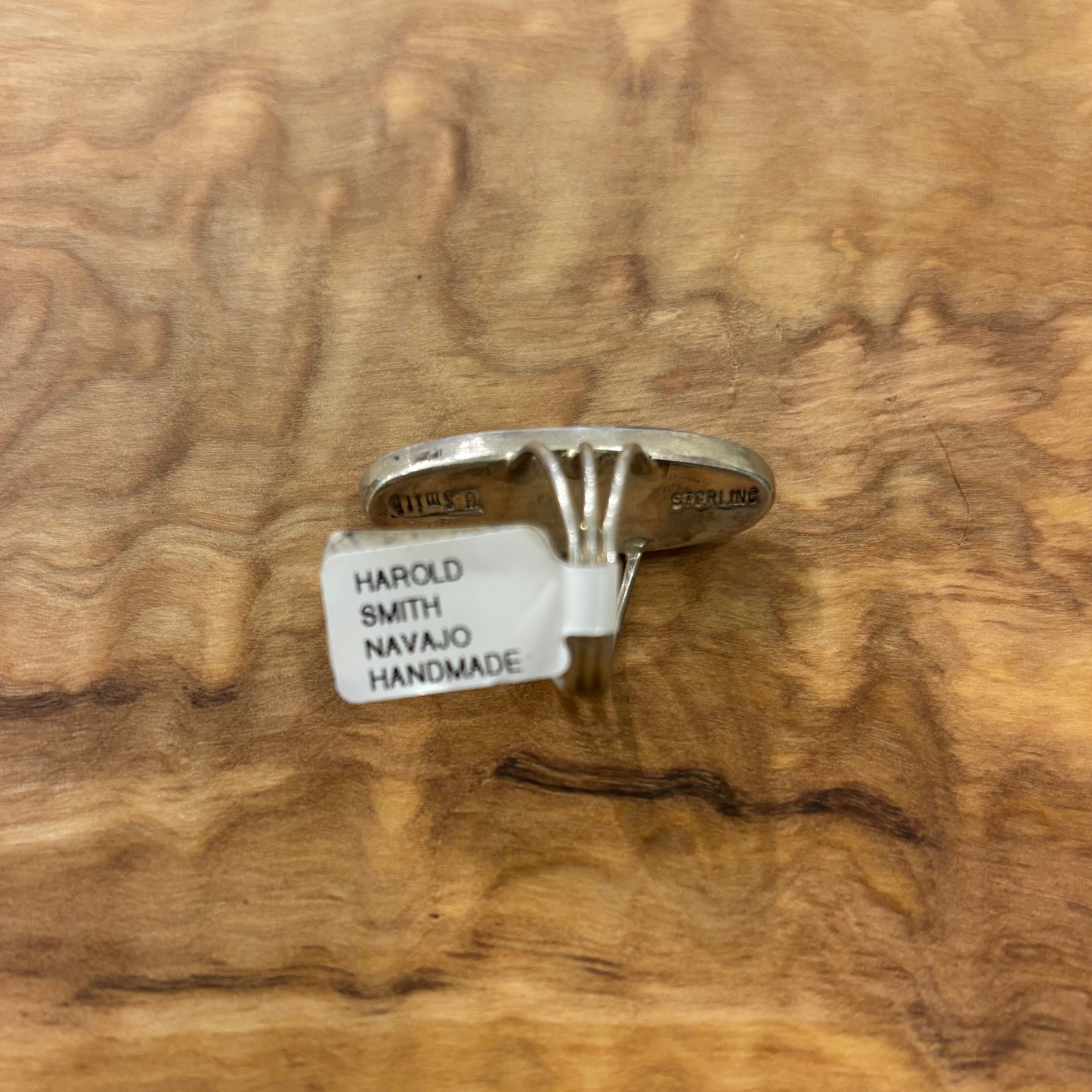 A sterling silver ring with a textured, sculpted band and a small white paper tag attached, displayed on a polished burl wood slab.